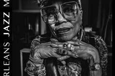 Black-and-white exhibition poster featuring a close-up portrait of a woman wearing artsy square glasses, layered bracelets, rings, and a patterned outfit, resting her chin on her hands. Large text reads “Less Is More: The Photography of Steve Rapport.” Vertical text along the side reads “New Orleans Jazz Museum.” The poster also includes details for an opening reception on April 21, 2026, at 6:00 p.m., featuring Joshua Starkman and friends.