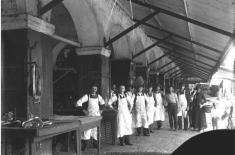 Black-and-white photograph of a row of butchers standing outside their stalls at the French Market in New Orleans. The men wear white aprons and stand beneath a covered arcade with arched openings and exposed beams. Meat hangs in the foreground, and a few women and a child stand farther down the walkway.