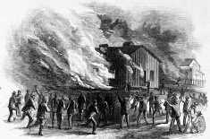 Black-and-white 19th-century-style engraving depicting a crowd of men gathered around a wooden building that is engulfed in flames. Thick smoke billows into the sky as the fire spreads. Several figures in the foreground raise their arms or gesture toward the blaze, while others run or stand watching. Additional structures appear in the background, suggesting a town or settlement. The scene conveys chaos and urgency as the fire consumes the building.