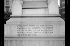 Black-and-white photograph of the base of a stone monument with an engraved inscription. The text states that U.S. troops took over the state government during Reconstruction and claims that the 1876 national election “recognized white supremacy in the South.” The carved message reflects a Lost Cause interpretation of events.