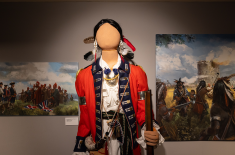 Mannequin dressed in an 18th-century military uniform with a red coat, blue trim, and decorative medals stands centered in a gallery. The figure wears feather adornments and holds a musket. Behind it, two large paintings depict scenes of colonial-era conflict, including soldiers and Indigenous warriors in battle near a fort under a cloudy sky.
