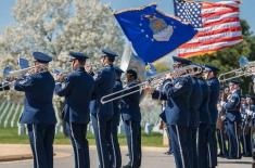 U.S. Air Force Band members in dress uniforms perform outdoors with brass instruments and drums. American and Air Force flags wave above the band, with rows of white headstones visible in the background.