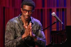 Kyle Roussel sits at a piano onstage beside a microphone, hands clasped, with warm red and purple lighting in the background.