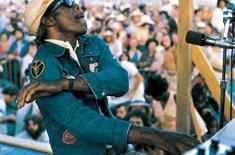 Professor Longhair performs at a piano on an outdoor stage, wearing sunglasses and a cap, with a large festival crowd in the background.
