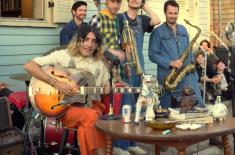 A band performs outdoors in front of a house, with a seated guitarist singing into a microphone while other musicians stand behind him holding brass and woodwind instruments.