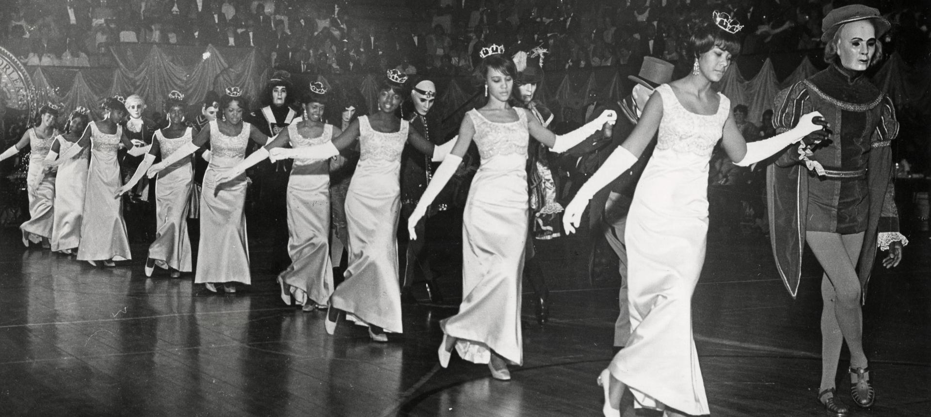 Black-and-white photograph of a line of women in gowns and tiaras, wearing long white gloves, dancing in formation with partners dressed in costumes. The audience is visible in the background watching the ball performance. Original Illinois Club debutantes and members dancing the Chicago Glide, 1968. Photograph by Porter’s Studio.