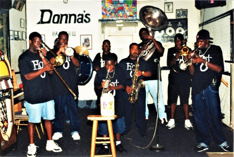 A group of seven musicians, including children, perform together indoors, playing brass and percussion instruments. A large “Donna’s” sign and framed artwork are visible on the wall behind them.  