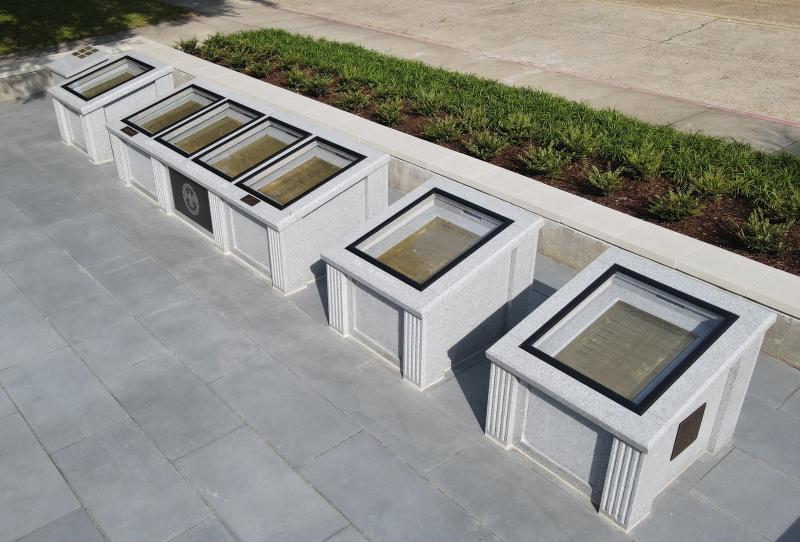 An aerial view of an outdoor exhibit featuring several granite displays arranged in a row on a grey stone patio. The displays, viewed from a high angle on a sunny day, are rectangular and made of light-colored, speckled granite. Each has a glass top protecting a golden plaque inscribed with text. Etched lettering on the sides of the displays identify the documents within, including the "United States Constitution," "Bill of Rights," and "Civil Rights Amendments." A bed of green shrubs and mulch runs alongsi