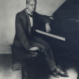 Black and white portrait of a man in a tuxedo seated at a piano bench, one arm resting on the piano keys. He faces the camera with a composed expression. Artifact from the exhibition "The District: The Music and Musicians of Storyville."