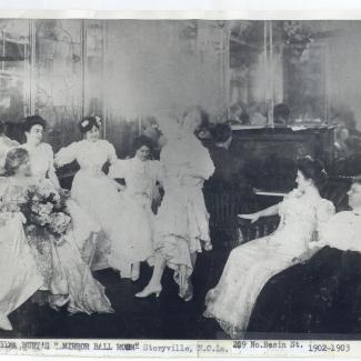 Black-and-white photograph of women in elegant early 1900s gowns gathered in a parlor known as “Emma Burt’s Mirror Ball Room” in Storyville, New Orleans, around 1902–1903. One woman dances while others sit and watch, surrounded by ornate mirrors, stained glass, and a piano in the background. The scene captures the social atmosphere of New Orleans’s Storyville era.