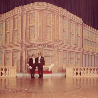 Color photograph of two men in tuxedos standing on a stage in front of a large painted backdrop depicting the French Opera House. The set includes a red carpet and white balustrades, giving the appearance of a grand entrance. Marcus Neustadter Jr. and Carl Duvigneaud at the Original Illinois Club ball, 1968. Photograph by Celeste Broadway.