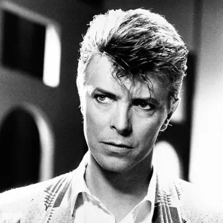 Black-and-white portrait of David Bowie looking intensely off camera, with styled hair, a white shirt, and a patterned jacket against a blurred urban background.