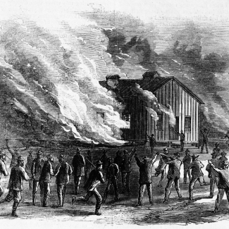 Black-and-white 19th-century-style engraving depicting a crowd of men gathered around a wooden building that is engulfed in flames. Thick smoke billows into the sky as the fire spreads. Several figures in the foreground raise their arms or gesture toward the blaze, while others run or stand watching. Additional structures appear in the background, suggesting a town or settlement. The scene conveys chaos and urgency as the fire consumes the building.