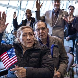 A group of people raise their right hands during a citizenship ceremony, with a woman in the foreground holding a small American flag.