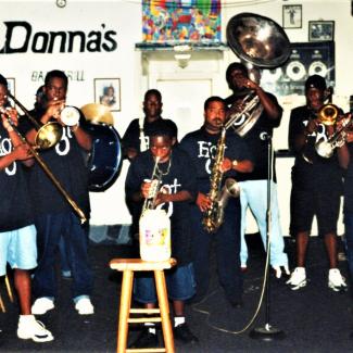 A group of seven musicians, including children, perform together indoors, playing brass and percussion instruments. A large “Donna’s” sign and framed artwork are visible on the wall behind them.  