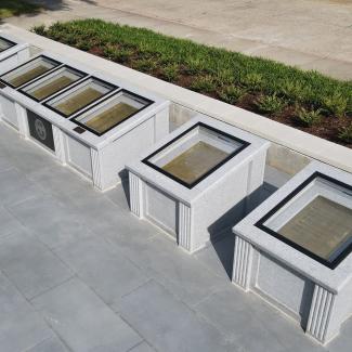 An aerial view of an outdoor exhibit featuring several granite displays arranged in a row on a grey stone patio. The displays, viewed from a high angle on a sunny day, are rectangular and made of light-colored, speckled granite. Each has a glass top protecting a golden plaque inscribed with text. Etched lettering on the sides of the displays identify the documents within, including the "United States Constitution," "Bill of Rights," and "Civil Rights Amendments." A bed of green shrubs and mulch runs alongsi